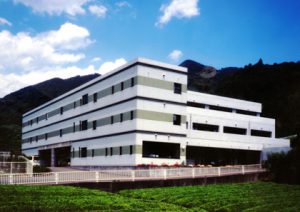 山間部に立つ白い4階建ての介護施設の建物外観。晴天で周辺は緑に囲まれている。